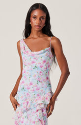 A woman is wearing a sleeveless, floral dress with a pastel color palette. The dress features thin spaghetti straps, a cowl neckline, and a ruffled hem detail. The fabric has a watercolor-style floral pattern in shades of pink, blue, green, and white.